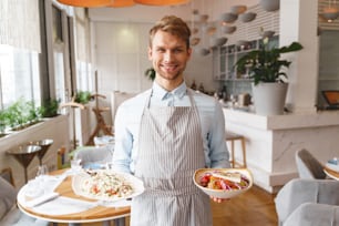 Handsome young man in apron looking at camera and smiling while holding delicious fresh salads