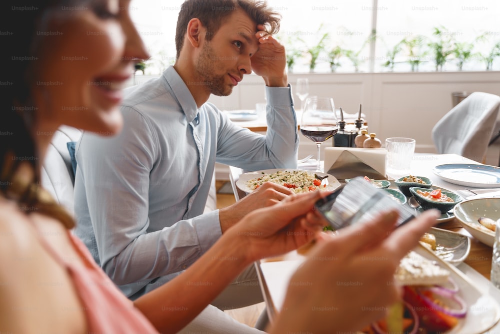 Charming lady talking food picture with smartphone while sitting at the table with good-looking gentleman