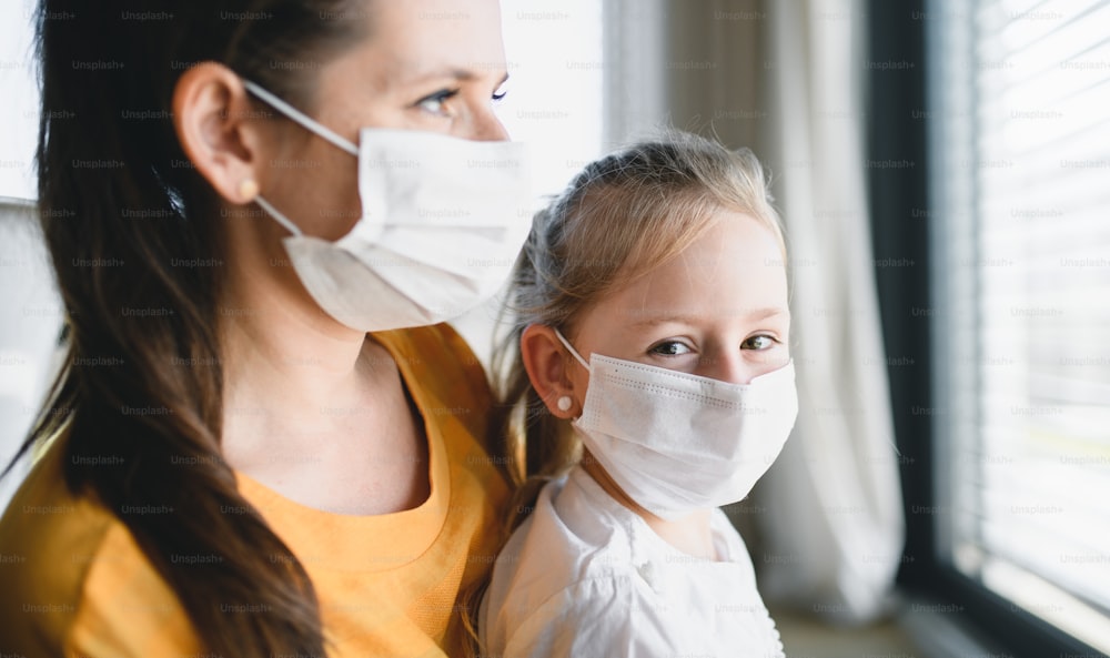 家の中でフェイスマスクをした母親と小さな子供が外を見ている。コロナウイルスと検疫のコンセプト。