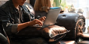 Foto de homem atraente em camisa jeans trabalhando / digitando em laptop de computador que colocando em seu colo enquanto sentado no sofá de couro sobre sala de estar confortável como fundo.