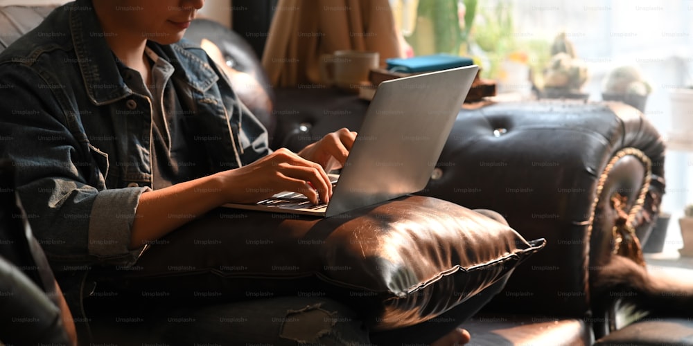 デニムシャツを着た魅力的な男性が、快適なリビングルームの上の革張りのソファに座りながら、膝の上に置いて、コンピューターのラップトップで作業/タイピングしている写真。