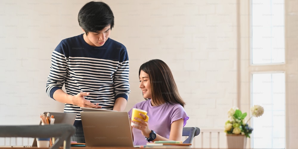 Photo of young couple working together with computer laptop and tablet while sitting and standing at the wooden working desk over modern sitting room as background.