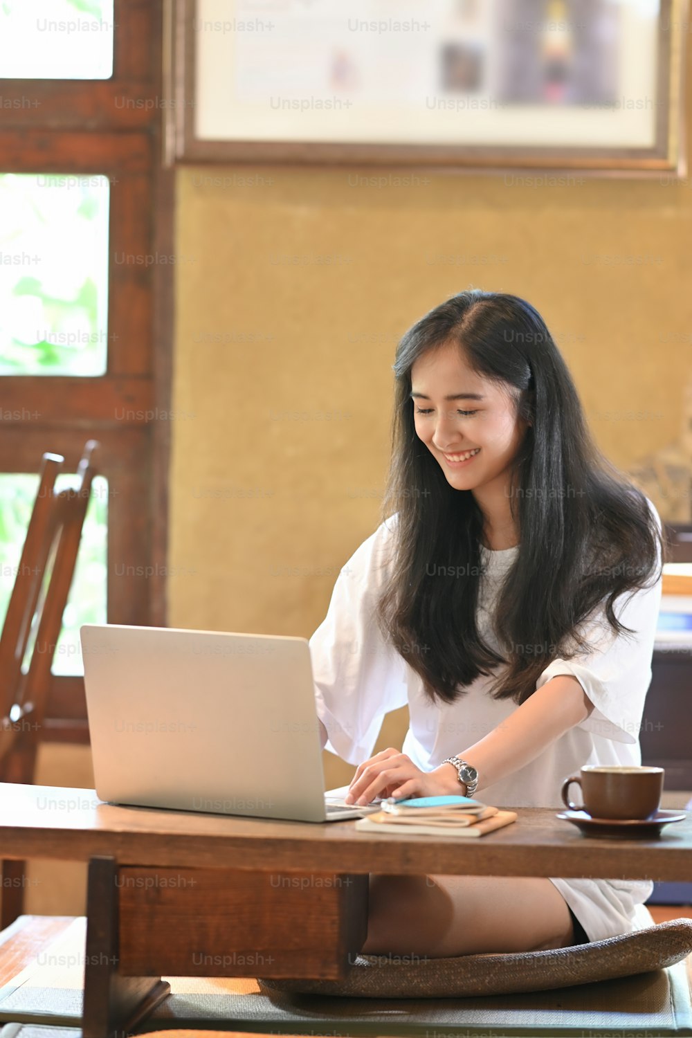 Photo of attractive asian woman working with computer laptop that putting on wooden working desk that surrounded by coffee cup and stack of notebook over vintage living room as background.