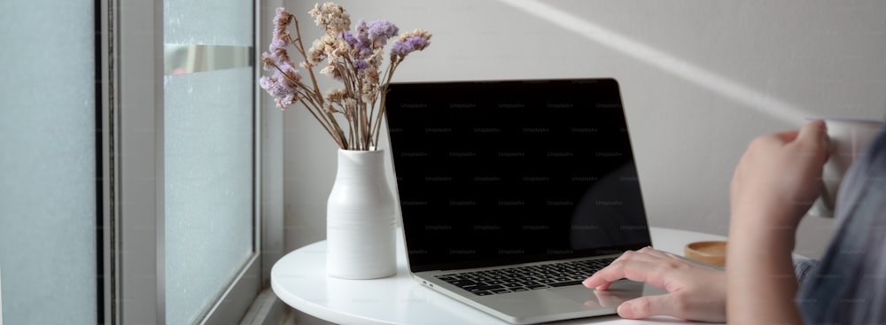 Cropped shot of female freelancer working on laptop with flower vase on white  circle coffee table at home