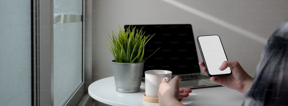 Colpo ritagliato di una freelance femminile che si rilassa con uno smartphone mock-up e un caffè caldo mentre lavora con il laptop nello spazio di lavoro portatile