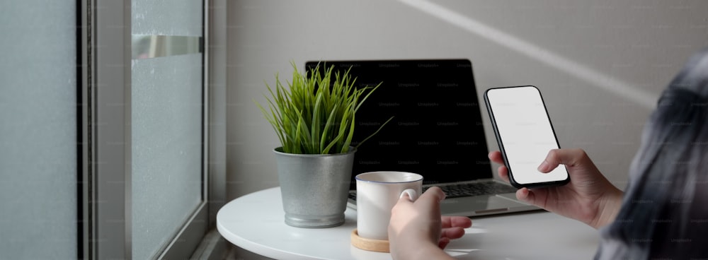 Ausschnittaufnahme einer Freiberuflerin, die sich mit einem nachgebauten Smartphone und heißem Kaffee entspannt, während sie mit dem Laptop am tragbaren Arbeitsplatz arbeitet