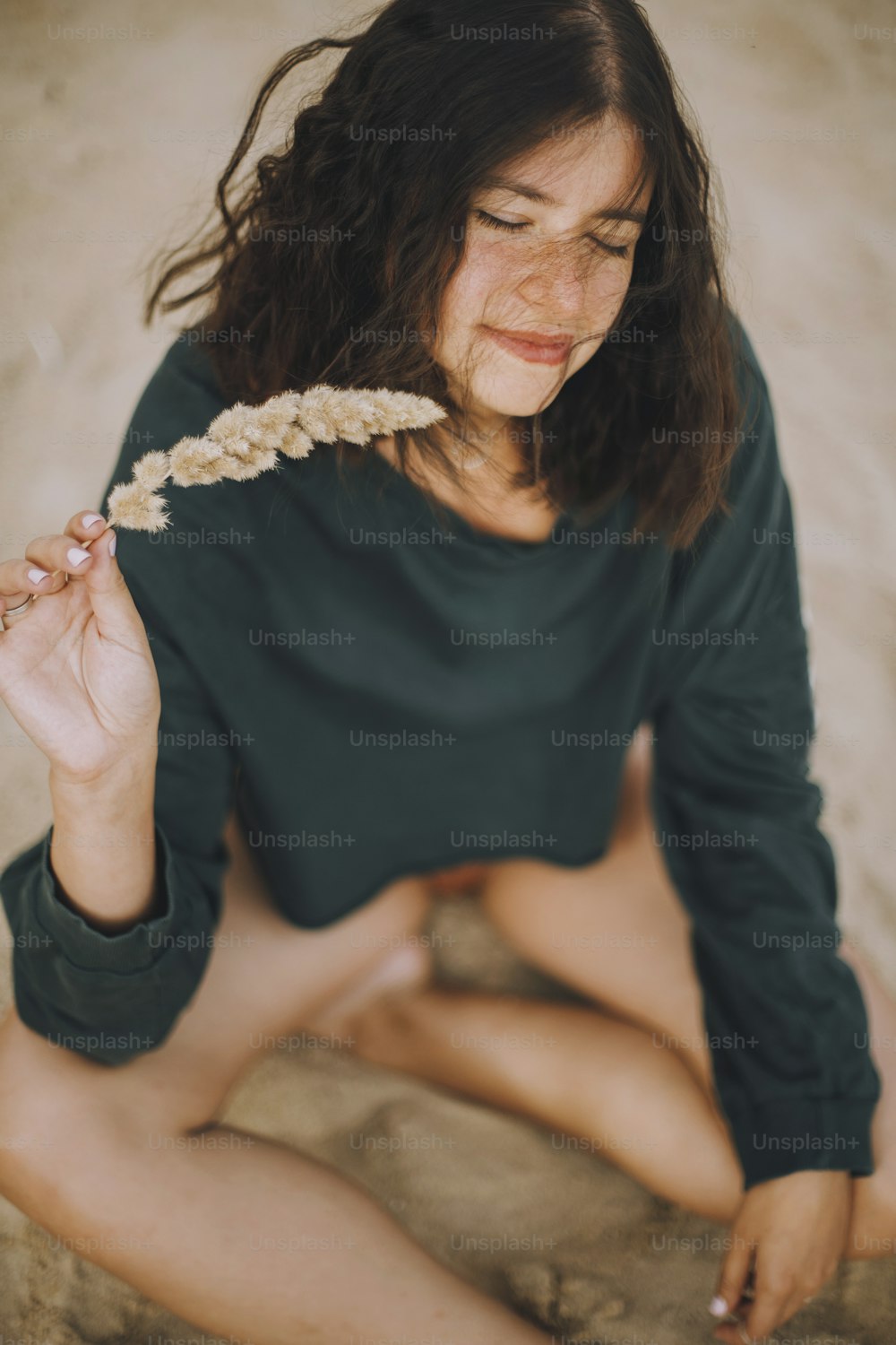 Elegante ragazza hipster che tiene l'erba e si siede sulla spiaggia sabbiosa. Giovane donna abbronzata alla moda in costume da bagno moderno e maglione che si rilassa in riva al mare. Vacanze estive. Spensierato
