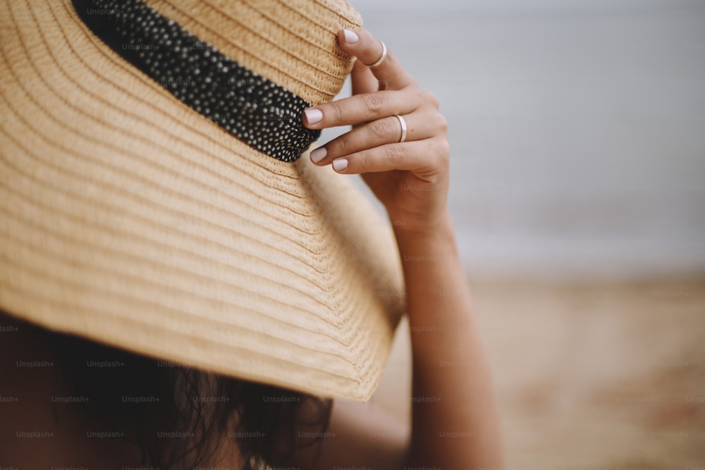 Vacaciones de verano y viajes. Muchacha sentada en la playa, mano sosteniendo sombrero de cerca. Mujer joven de moda con sombrero de paja relajándose en la playa de arena cerca del mar. Despreocupado. Espacio para el texto