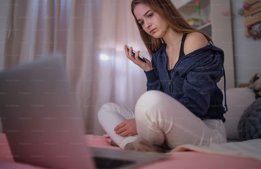 Worried young girl with smartphone sitting indoors on bed, online dating and internet abuse concept.
