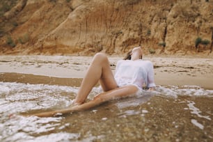 Giovane donna in camicia bianca seduta sulla spiaggia in onde che spruzzino. Elegante ragazza abbronzata che si rilassa in riva al mare e si gode le onde. Vacanze estive. Mindfulness e momento di spensieratezza