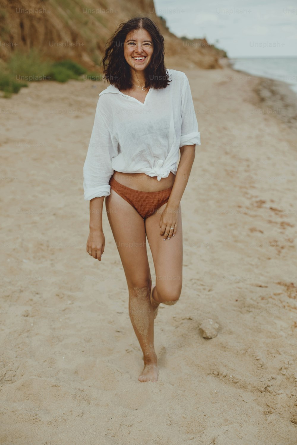 Glückliches Boho-Mädchen im weißen Hemd am sonnigen Strand spazieren gehend. Unbeschwerte stilvolle Frau in Badeanzug und Hemd entspannt sich am Meer. Sommerferien. Authentisches Lifestyle-Image
