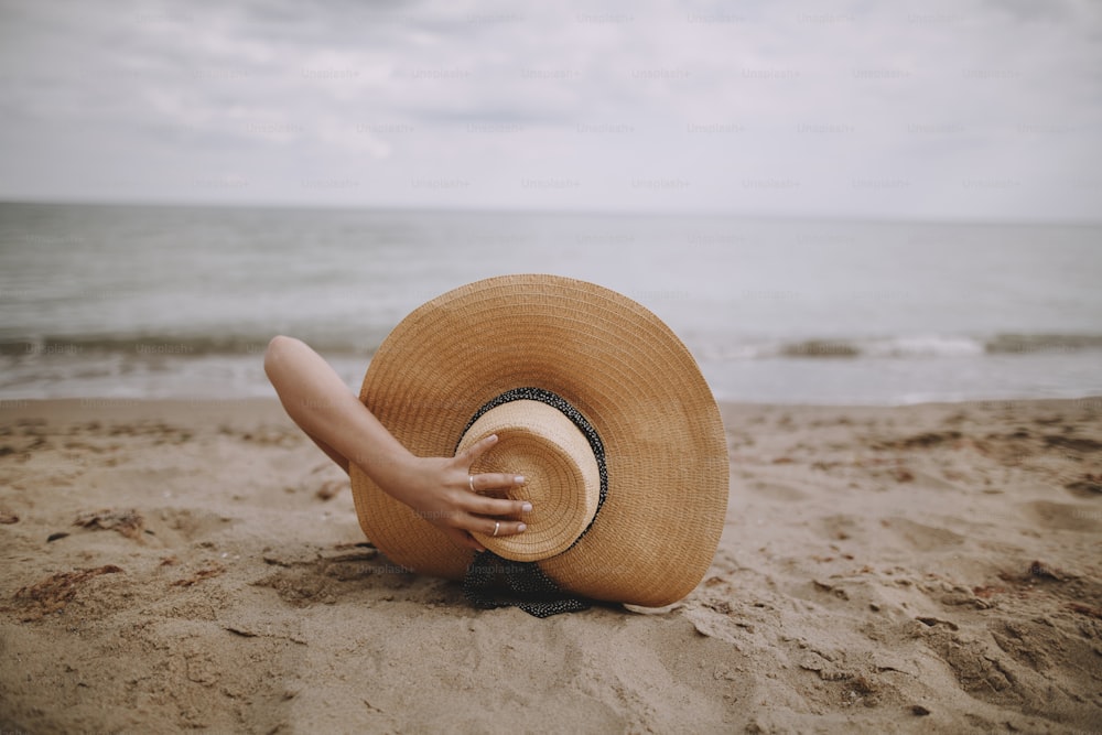 Summer vacation and travel. Girl in hat lying on beach. Fashionable young woman holding straw hat, relaxing on sandy beach near sea. Mindfulness and carefree creative image