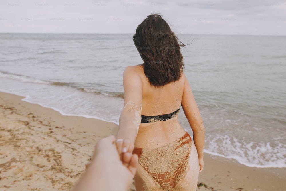 따라 오세요. 해변에서 손을 잡고 남자를 이끄는 여자. 여름 방학이나 신혼 여행에 사랑에 빠진 커플. 남자의 손을 잡고 있는 모래 검게 그을린 몸을 가진 젊은 여자