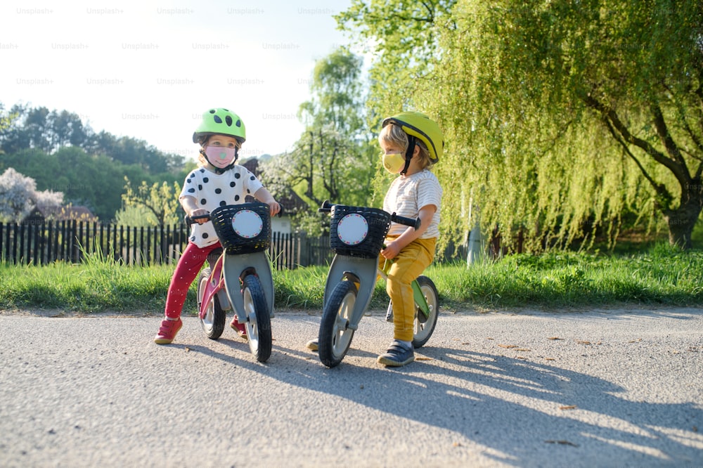 Small children boy and girl with face masks playing outdoors with bike, coronavirus concept.