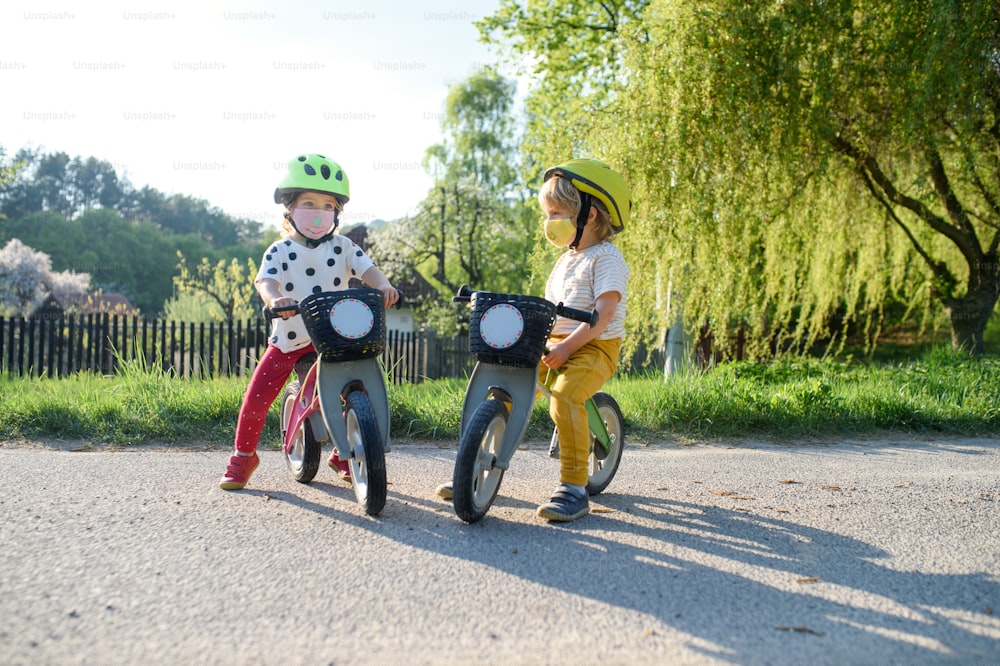 Crianças pequenas menino e menina com máscaras faciais brincando ao ar livre com bicicleta, conceito de coronavírus.