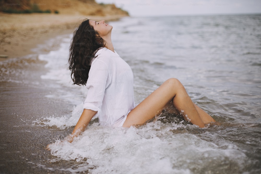 Happy young woman in white shirt sitting on beach in splashing waves. Stylish tanned girl relaxing on seashore and enjoying waves. Summer vacation. Mindfulness and carefree moment