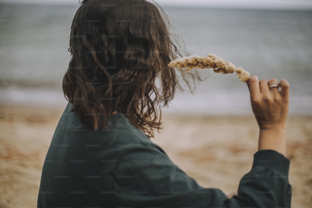 Fille hipster insouciante aux cheveux venteux tenant de l’herbe sur la plage. Femme bohème bronzée élégante dans une tenue moderne se relaxant au bord de la mer. Vacances. Consciencieux