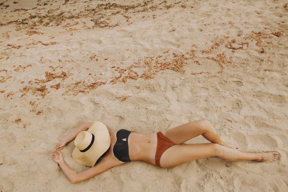 Fashionable young woman in straw hat relaxing on sandy beach near sea. Summer vacation and travel. Girl lying on beach with hand holding hat. Carefree. Top view