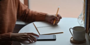 Imagen recortada de una mujer atractiva tomando notas mientras está sentada en el escritorio de trabajo blanco sobre una cómoda sala de estar como fondo.
