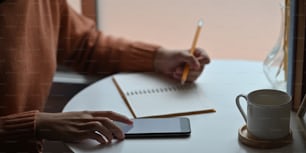 Cropped image of attractive woman taking notes while sitting at the white working desk over comfortable living room as background.