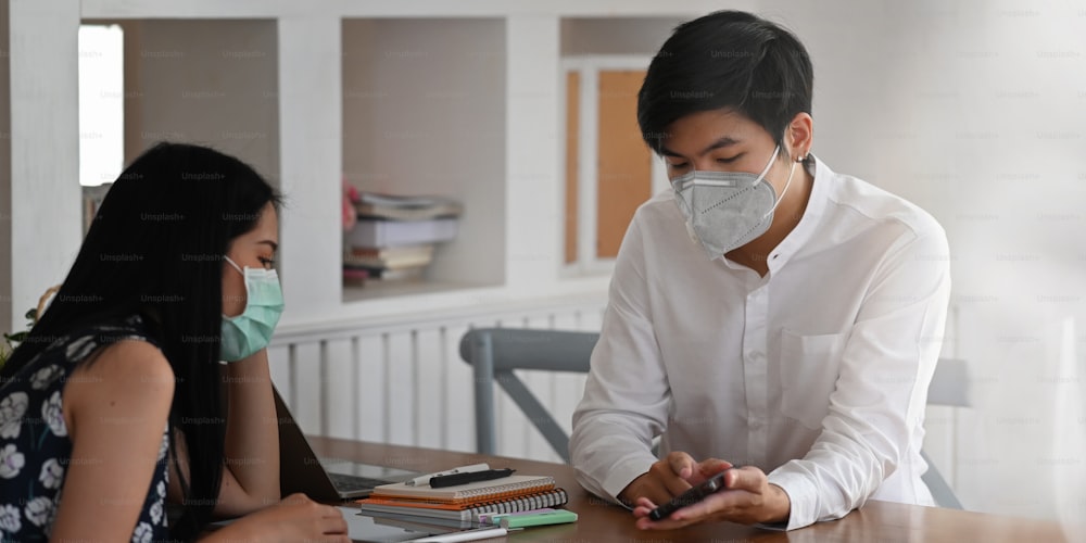 Photo of young couple that wearing a medical mask sitting and relaxing together at the wooden working desk over comfortable living room as background.