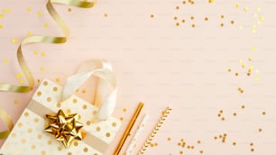 Golden party decorations on beige background top view. Flat lay shopping bag with gift, drinking straws, golden ribbon and confetti. Christmas, birthday or wedding concept