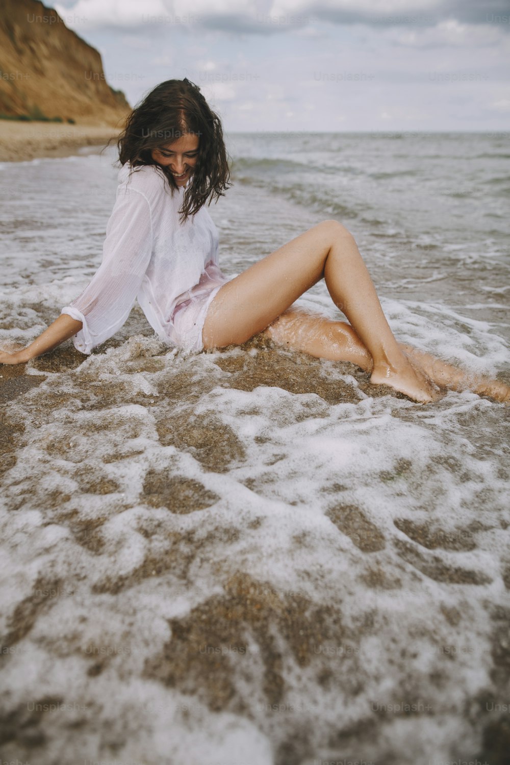Glückliche Frau im weißen Hemd sitzt am Strand in plätschernden Wellen. Stilvolles gebräuntes Mädchen, das sich am Meer entspannt und Wellen genießt. Sommerferien. Achtsamkeit und unbeschwerter Moment