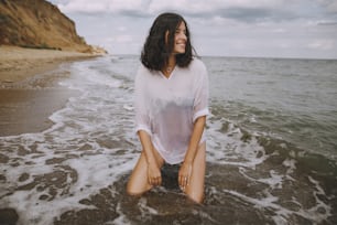 Young woman in white shirt sitting on beach in splashing waves. Stylish tanned girl relaxing on seashore and enjoying waves. Summer vacation. Mindfulness and carefree moment
