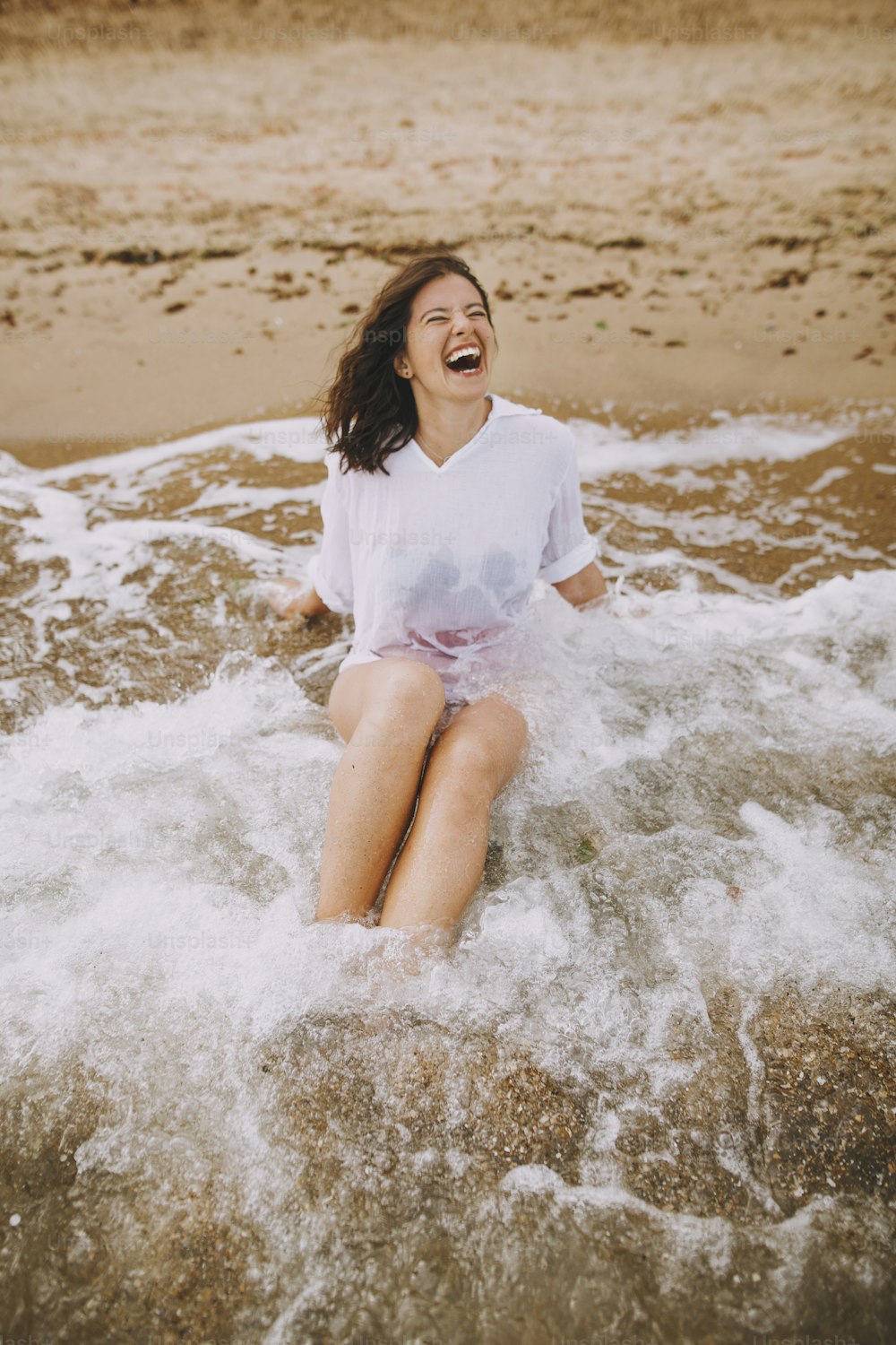 Happy young woman in wet white shirt lying on beach in splashing waves.  Emotional tanned girl relaxing on seashore and enjoying waves. Summer vacation. Carefree moment