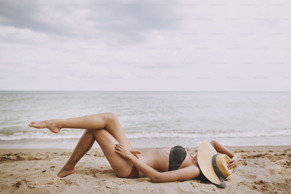 Stylish tanned girl in hat lying on beach. Fashionable young woman covering with straw hat, relaxing on sandy beach near sea. Summer vacation and travel on tropical island
