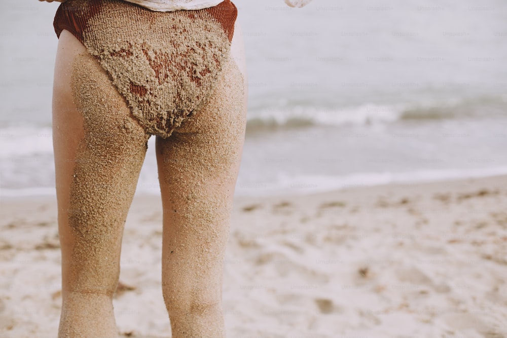 Bunda da mulher nova e pernas na areia fecham na praia. Menina molhada feliz com maiô de areia e pernas relaxando à beira-mar, vista para trás. Férias de verão. Momento autêntico despreocupado.