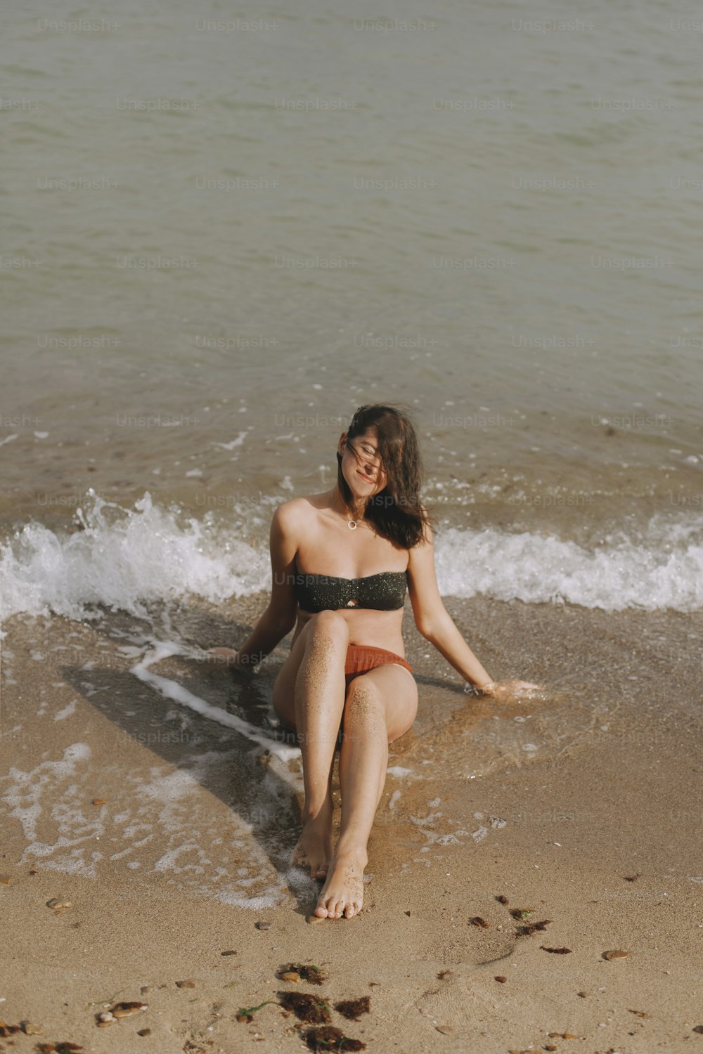 Joven feliz sentada en la playa con olas. Elegante chica bronceada en traje de baño moderno relajándose en la orilla del mar. Veraneo. Momento despreocupado