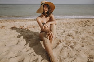 Stylish tan girl in hat sitting on beach. Fashionable young woman in straw hat, relaxing on sandy beach near sea in sunny light. Summer vacation and travel on tropical island
