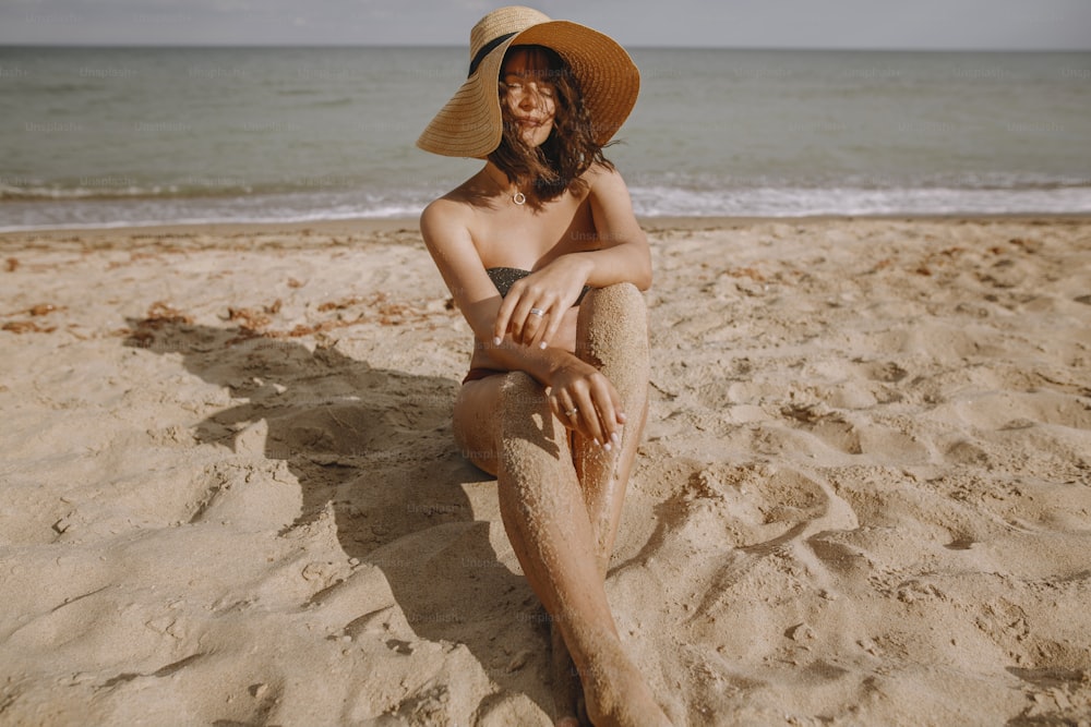 Elegante chica bronceada con sombrero sentada en la playa. Mujer joven de moda con sombrero de paja, relajándose en la playa de arena cerca del mar a la luz del sol. Vacaciones de verano y viajes en una isla tropical