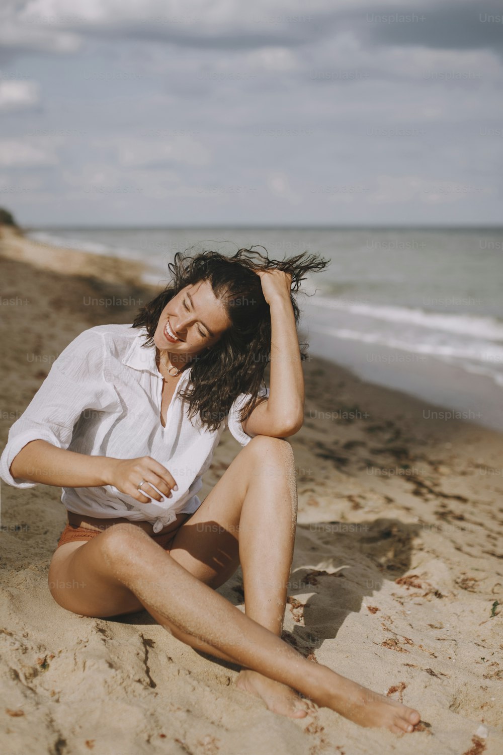 Joyful boho girl in white shirt sitting on sunny beach. Carefree stylish woman smiling and relaxing on seashore. Summer vacation. Lifestyle authentic image. Mindfulness