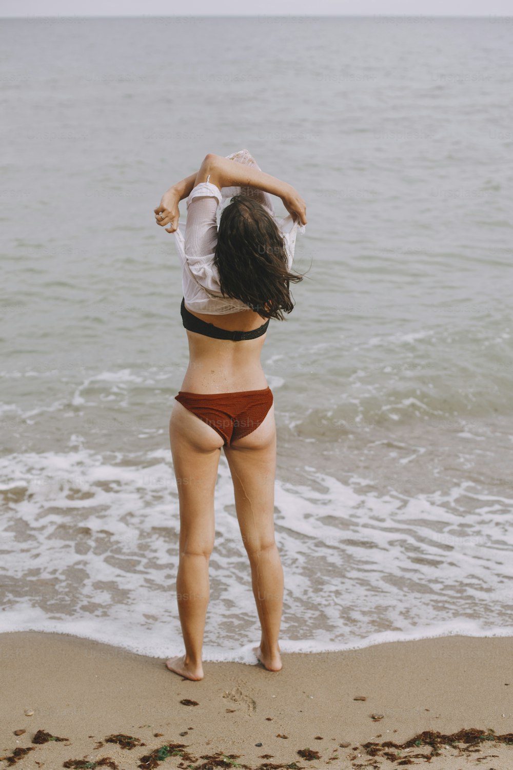 Mujer joven feliz quitándose la camisa blanca mojada en la playa, vista trasera. Chica elegante en traje de baño relajándose en la orilla del mar. Veraneo. Momento despreocupado