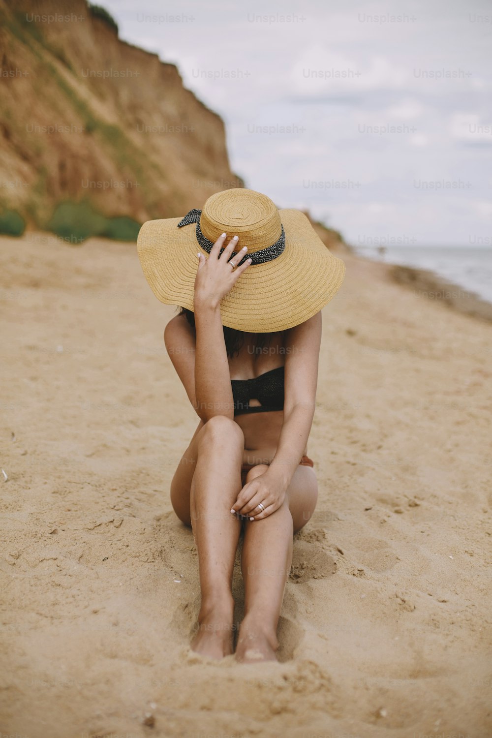 Stylish tanned girl in hat sitting on beach. Fashionable young woman covering with straw hat, relaxing on sandy beach near sea. Summer vacation and travel on tropical island