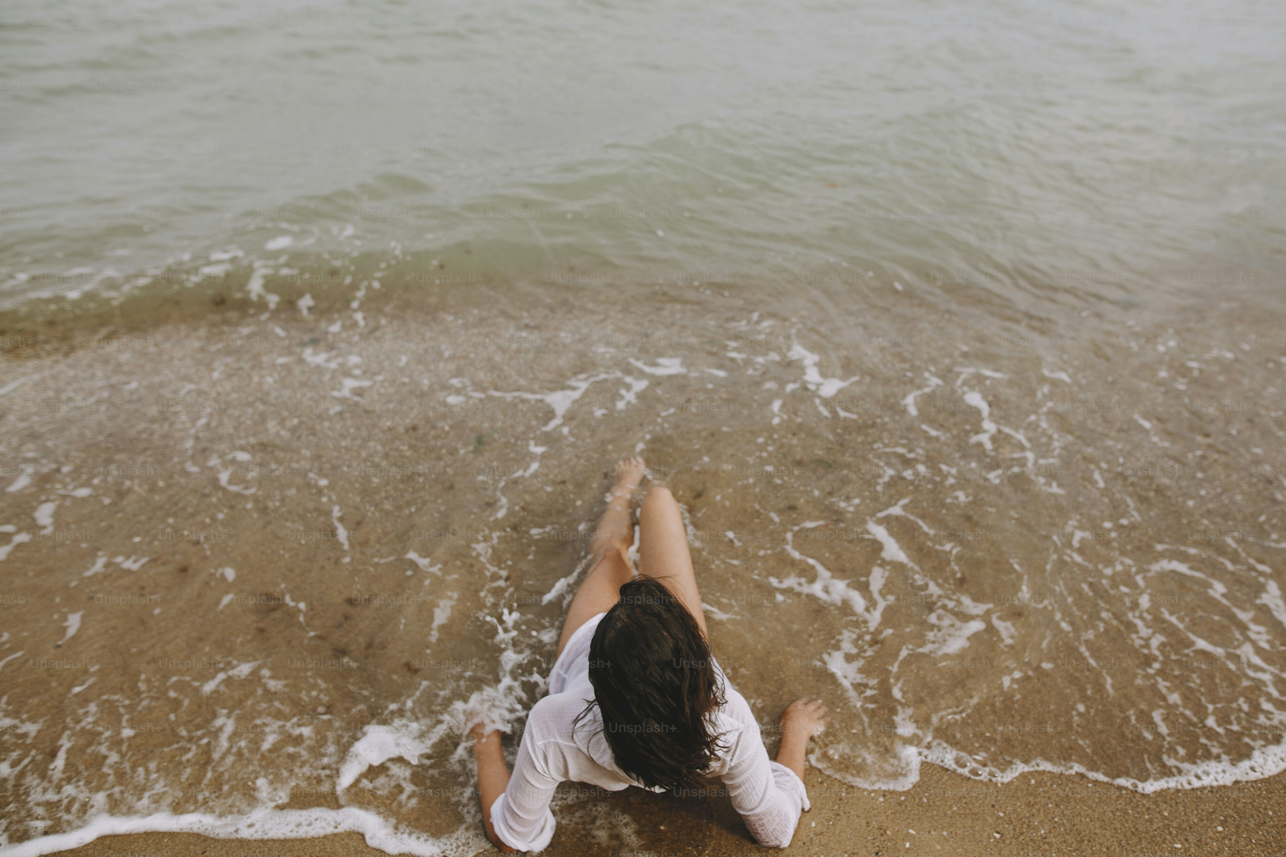 Mujer joven con camisa blanca mojada que yace en la playa en olas que salpican. Vista superior. Elegante chica bronceada relajándose en la orilla del mar y disfrutando de las olas. Veraneo. Mindfulness y despreocupación