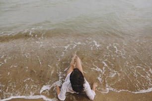 Young woman in wet white shirt lying on beach in splashing waves. Top view. Stylish tanned girl relaxing on seashore and enjoying waves. Summer vacation. Mindfulness and carefree