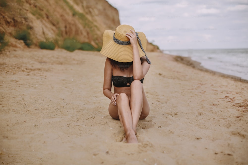 Stylish tanned girl in hat sitting on beach. Fashionable young woman covering with straw hat, relaxing on sandy beach near sea. Summer vacation and travel on tropical island
