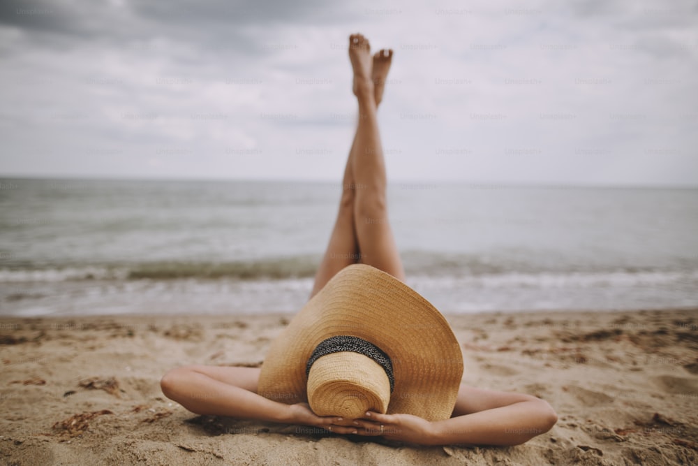 Summer vacation and travel. Girl in hat lying on beach with legs up. Fashionable young woman holding straw hat, relaxing on sandy beach near sea. Carefree creative image