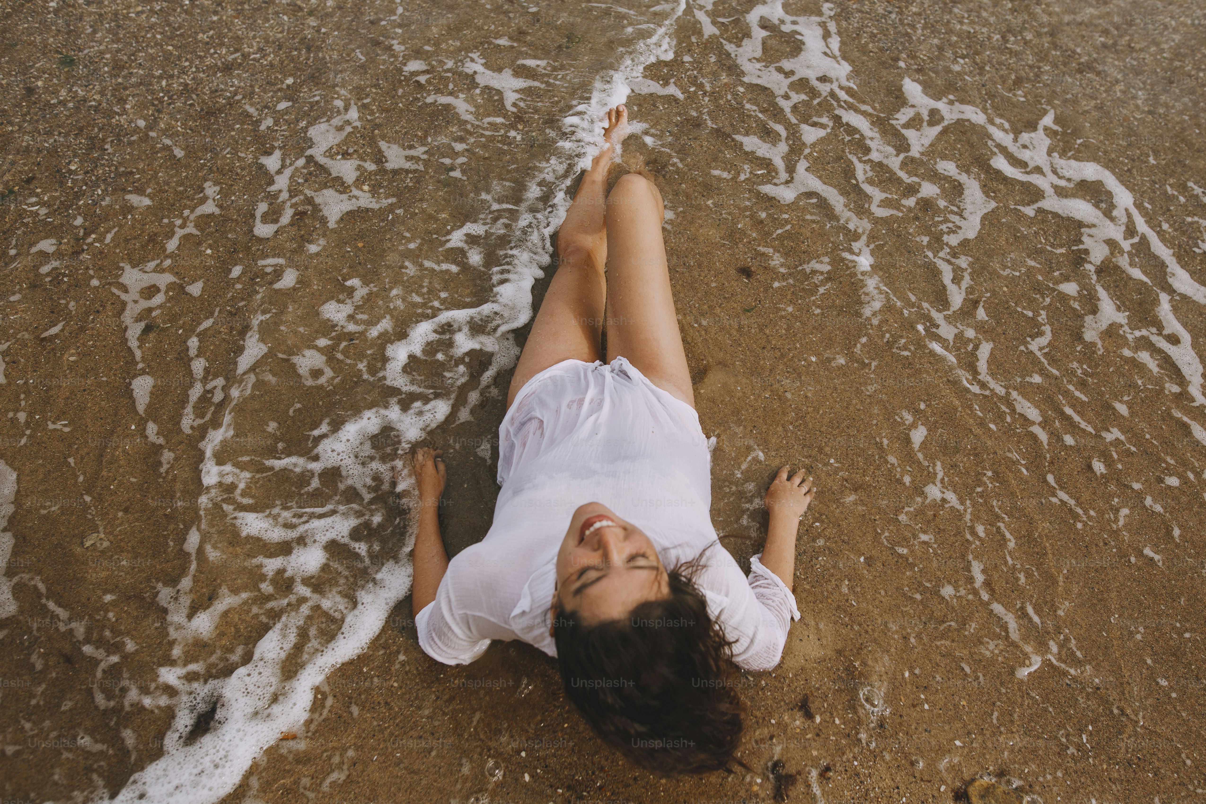 Mujer joven feliz con camisa blanca acostada en la playa en olas que salpican. Vista superior. Elegante chica bronceada relajándose en la orilla del mar y disfrutando de las olas. Veraneo. Mindfulness y despreocupación