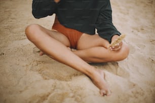 Hipster girl holding herb and sitting on beach, cropped view on tanned legs. Stylish boho woman in modern swimsuit and sweater relaxing on seashore. Summer vacation. Mindfulness