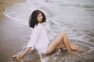 Mujer joven feliz con camisa blanca sentada en la playa en olas que salpican. Elegante chica bronceada relajándose en la orilla del mar y disfrutando de las olas. Veraneo. Mindfulness y momento despreocupado