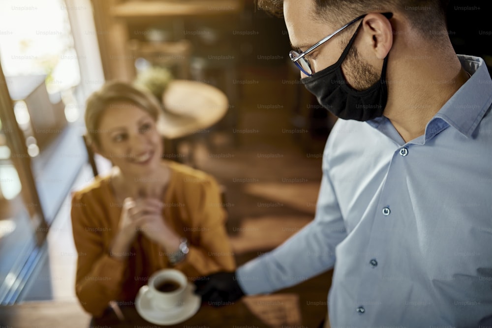 Young waiter serving coffee to a customer while wearing protective face mask in a cafe.