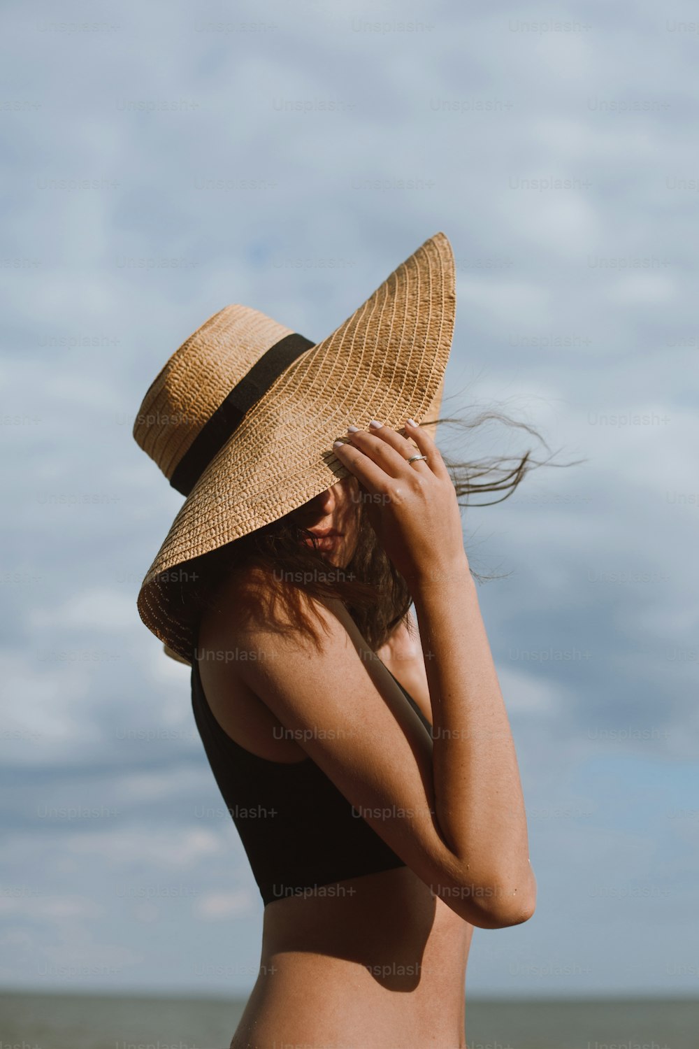 Menina sensual despreocupada segurando chapéu e posando no fundo do céu azul no dia quente do verão. Mulher bronzeada elegante no chapéu de palha relaxando na praia. Férias de verão