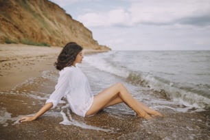 Happy young woman in white shirt sitting on beach in splashing waves. Stylish tanned girl relaxing on seashore and enjoying waves. Summer vacation. Mindfulness and carefree moment