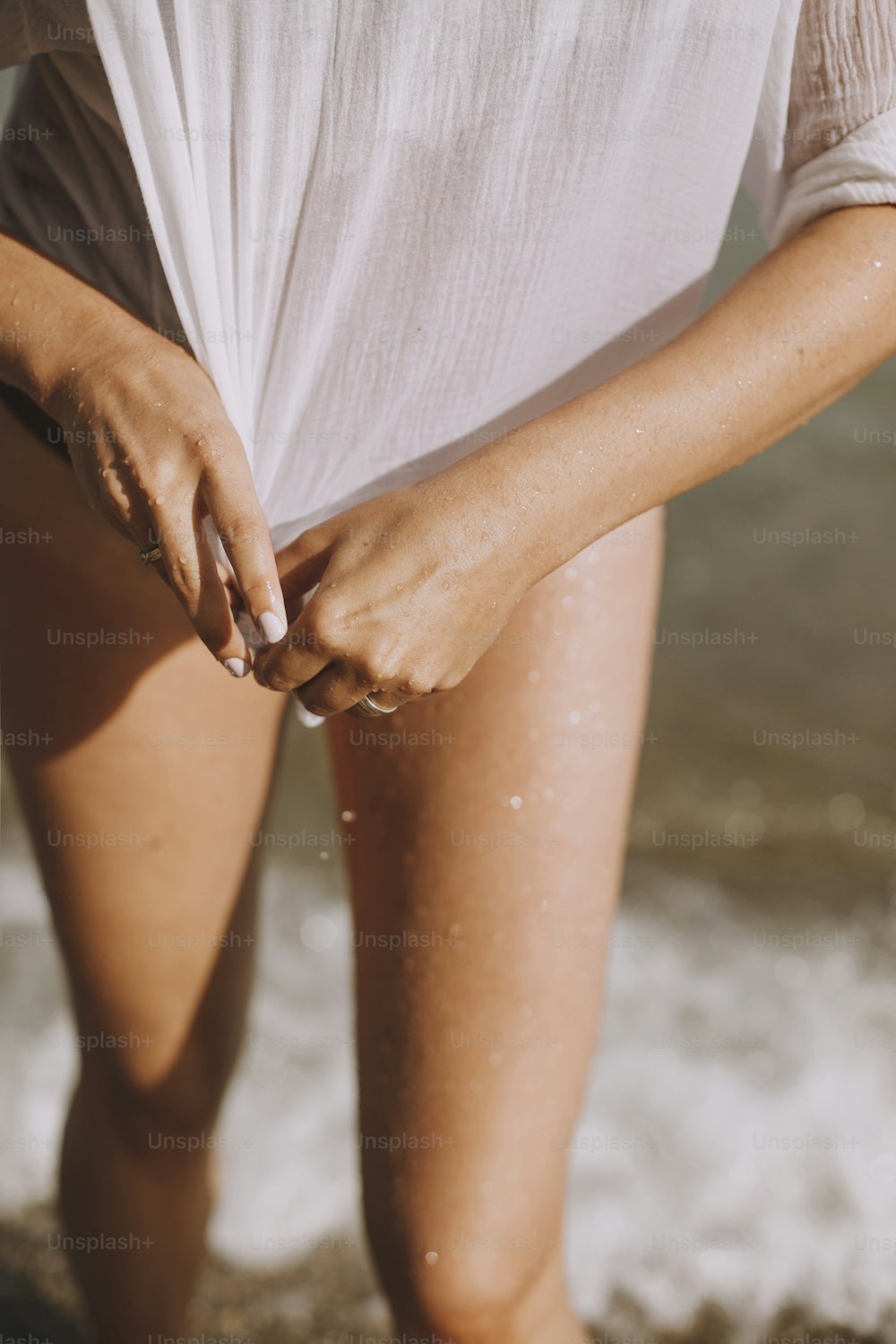 Young woman holding wet white shirt in hands on beach. Stylish tanned girl relaxing on seashore in waves. Summer vacation. Carefree funny moment
