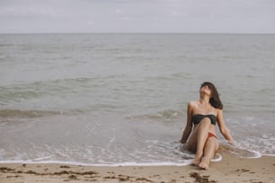 Giovane donna felice seduta sulla spiaggia con le onde. Elegante ragazza abbronzata in costume da bagno moderno che si rilassa in riva al mare. Vacanze estive. Momento spensierato. Immagine autentica