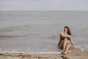 Happy young woman sitting on beach with waves. Stylish tanned  girl in modern swimsuit relaxing on seashore. Summer vacation. Carefree moment. Authentic image